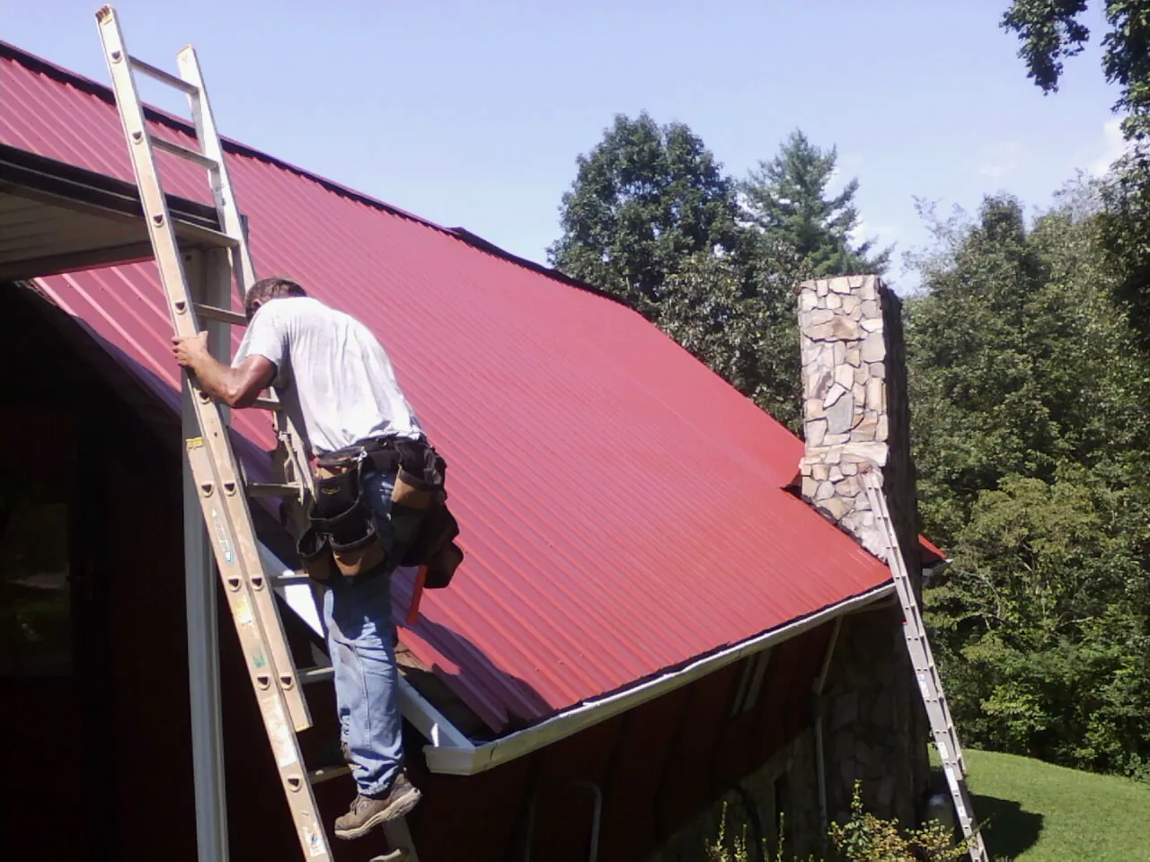 Roofer on a ladder beside a red metal roof and stone chimney — Soffit & Fascia Repair by Roofing Brothers in Pleasureville